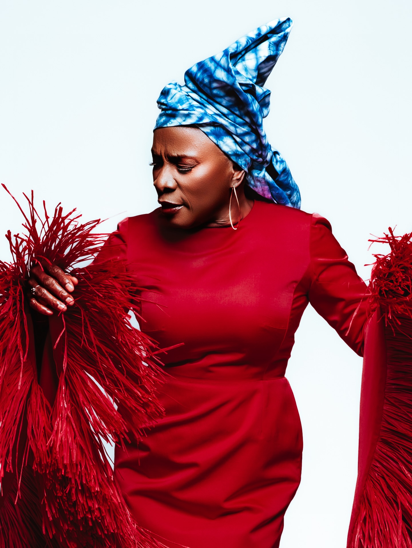 Angelique Kidjo holding the layered red raffia fringe sleeve of her crimson Imane Ayissi gown, wearing a structured sky-blue tie-dye headwrap and thin gold hoop earrings.