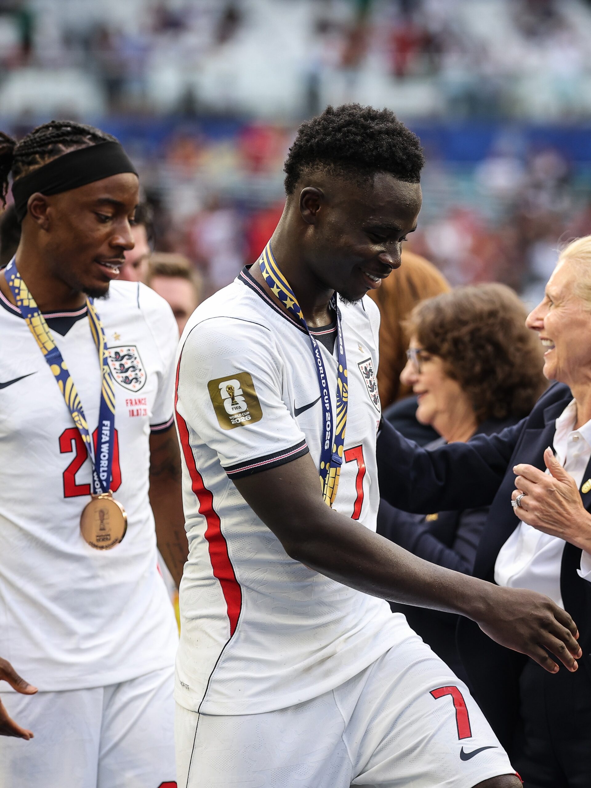 Bukayo Saka smiles whilst wearing his FIFA World Cup 2026 bronze medal on the pitch next to teammate Anthony Gordon.