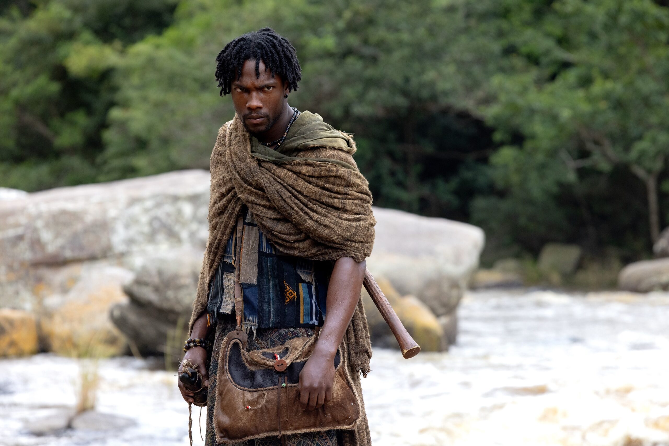 Tosin Cole as Tzain wearing a textured brown shawl and patterned trousers, standing on a rocky riverbank in Orïsha.