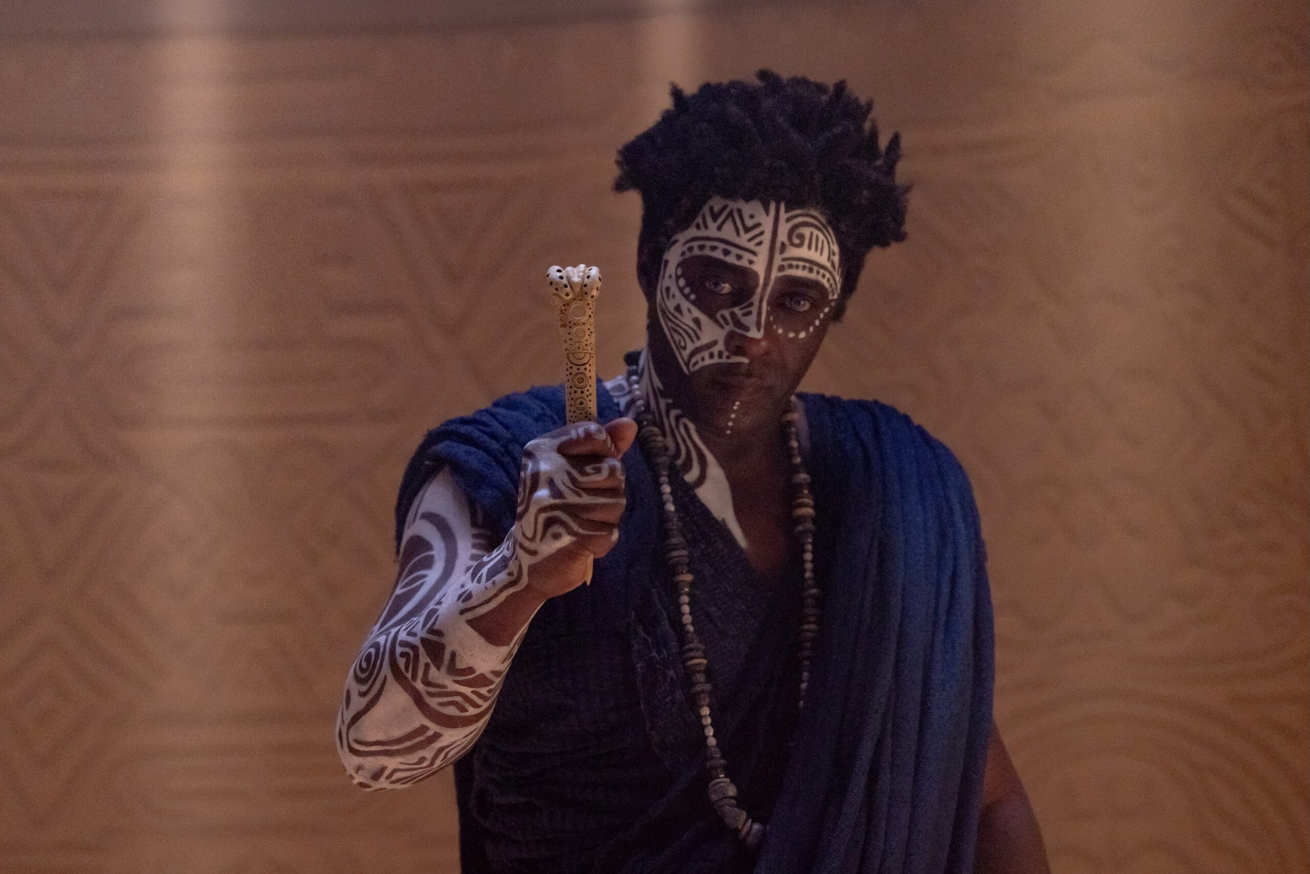Damson Idris as Prince Inan wearing a blue wrap with white traditional facial paint and body art, holding a carved sceptre.