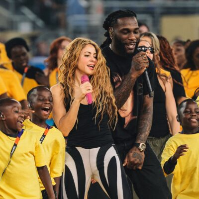 Shakira sings into a pink microphone while Burna Boy holds a black microphone during a stage rehearsal surrounded by dancing children in yellow shirts.