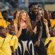 Shakira sings into a pink microphone while Burna Boy holds a black microphone during a stage rehearsal surrounded by dancing children in yellow shirts.
