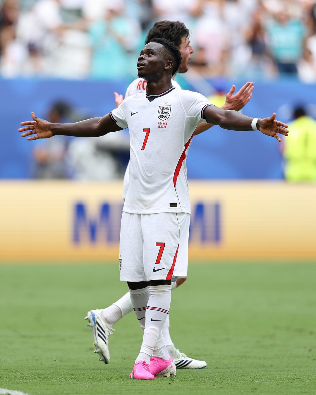 Bukayo Saka stands on the pitch looking upwards with his arms spread wide open following a goal against France.