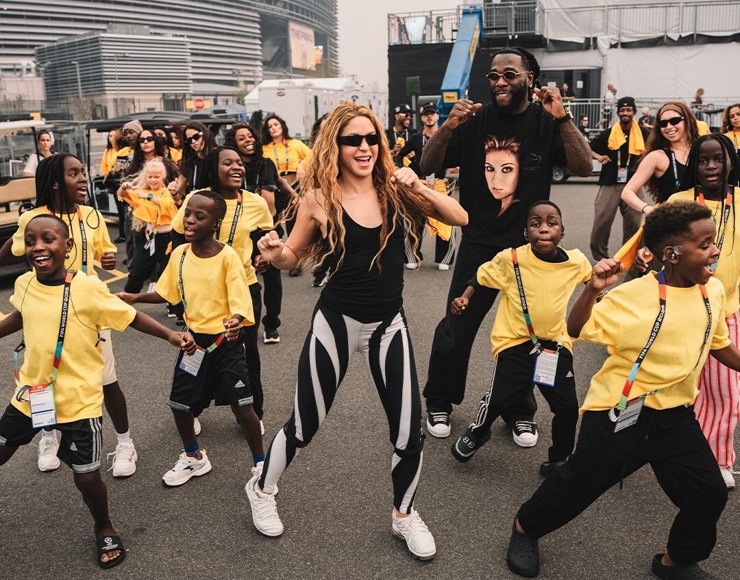 Shakira and Burna Boy dance outdoors alongside the Ghetto Kids of Uganda wearing yellow t-shirts during FIFA World Cup halftime rehearsals.