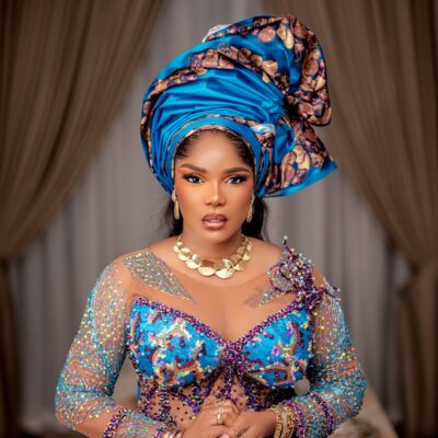 Iyabo Ojo in a detailed turquoise and purple illusion corset gown, showcasing the structured metallic gele and gold choker necklace.