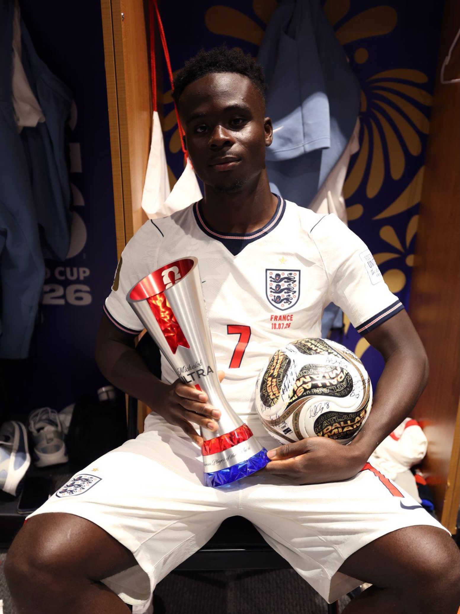 Bukayo Saka in the stadium dressing room holding the official Man of the Match trophy and the signed match ball.