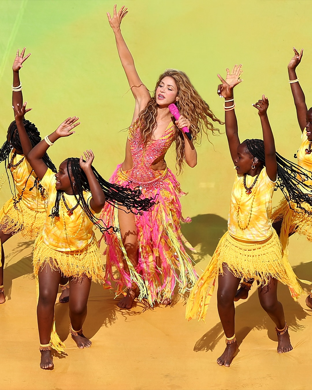 Shakira in a pink and yellow beaded bodysuit sings into a pink microphone alongside Uganda's Ghetto Kids dancing in yellow tie-dye outfits.
