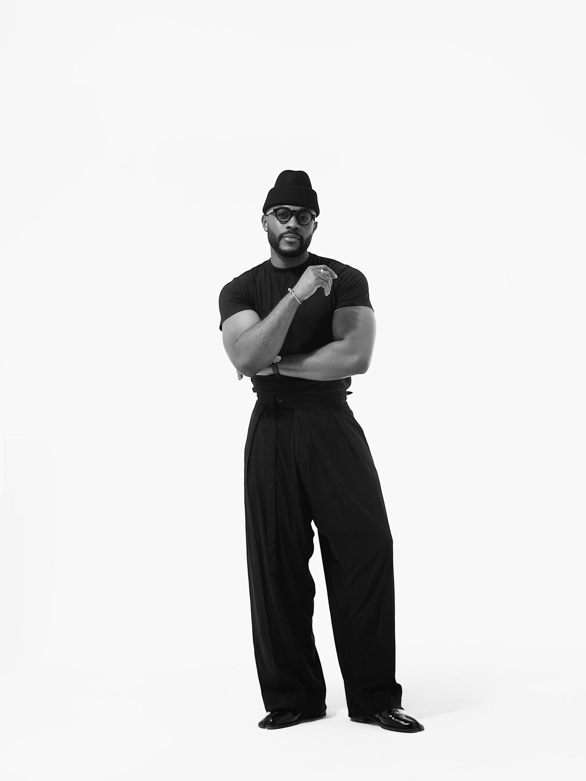 Kola Omotosho wearing a black t-shirt, high-waisted sash trousers, black beanie, and thick-rimmed glasses styled by Yomo Daj.