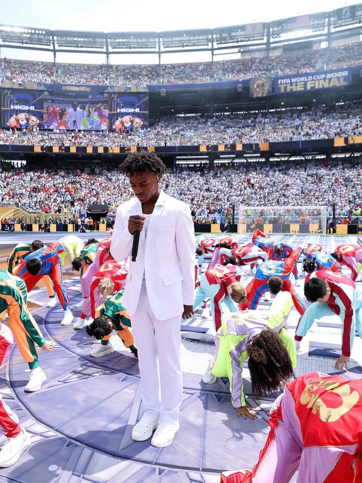 IShowSpeed in a white suit holding a microphone on the pitch surrounded by dancers at MetLife Stadium.