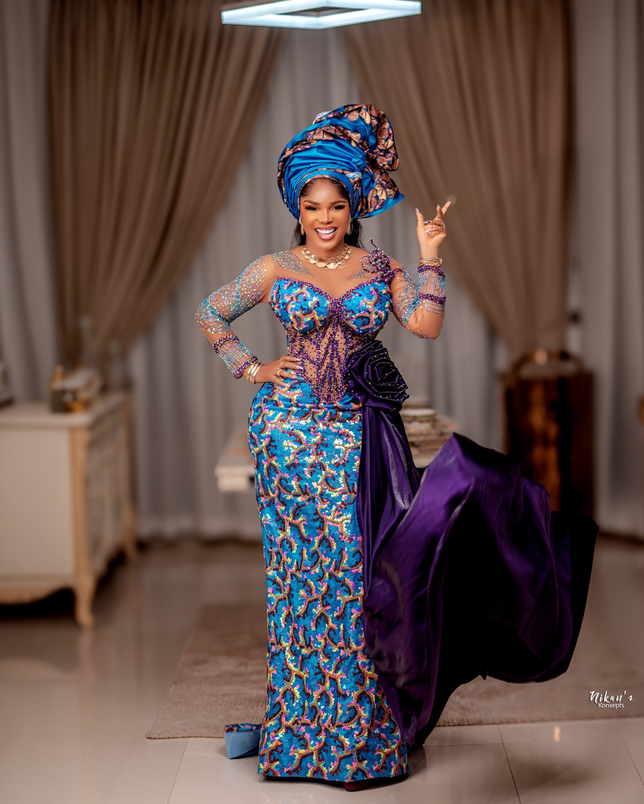 Iyabo Ojo posing to show the fitted silhouette of the turquoise lace gown, highlighting the deep purple velvet rosette and sweeping satin train.