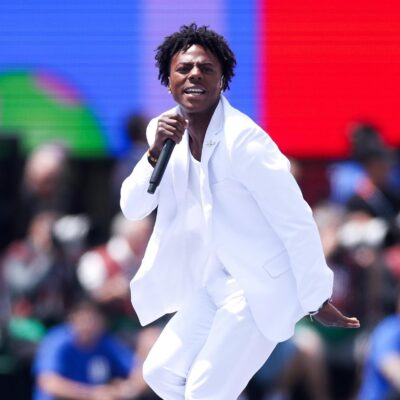 IShowSpeed dancing and singing into a microphone in an all-white suit on stage.