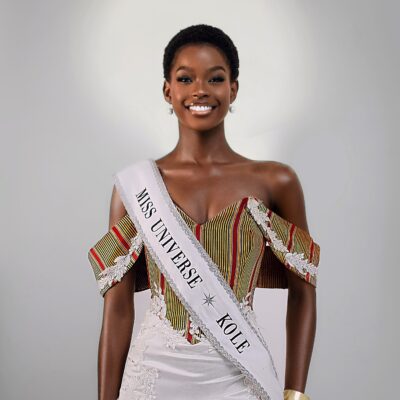 Kimberly Jael Aremo Acio wearing the official Miss Universe Kole sash over a white lace-embellished gown with an off-the-shoulder striped corset top for her official pageant delegate portrait.