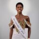 Kimberly Jael Aremo Acio wearing the official Miss Universe Kole sash over a white lace-embellished gown with an off-the-shoulder striped corset top for her official pageant delegate portrait.