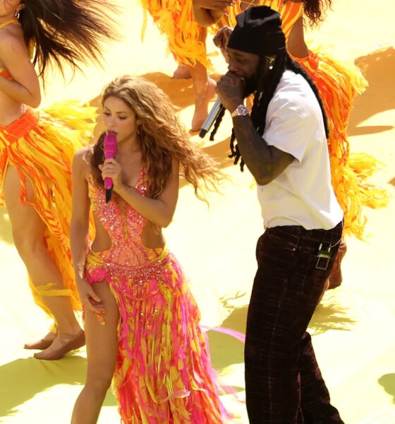 Burna Boy in a white t-shirt, dark trousers, and black beanie sings into a silver microphone beside Shakira performing barefoot on stage.