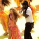 Burna Boy in a white t-shirt, dark trousers, and black beanie sings into a silver microphone beside Shakira performing barefoot on stage.