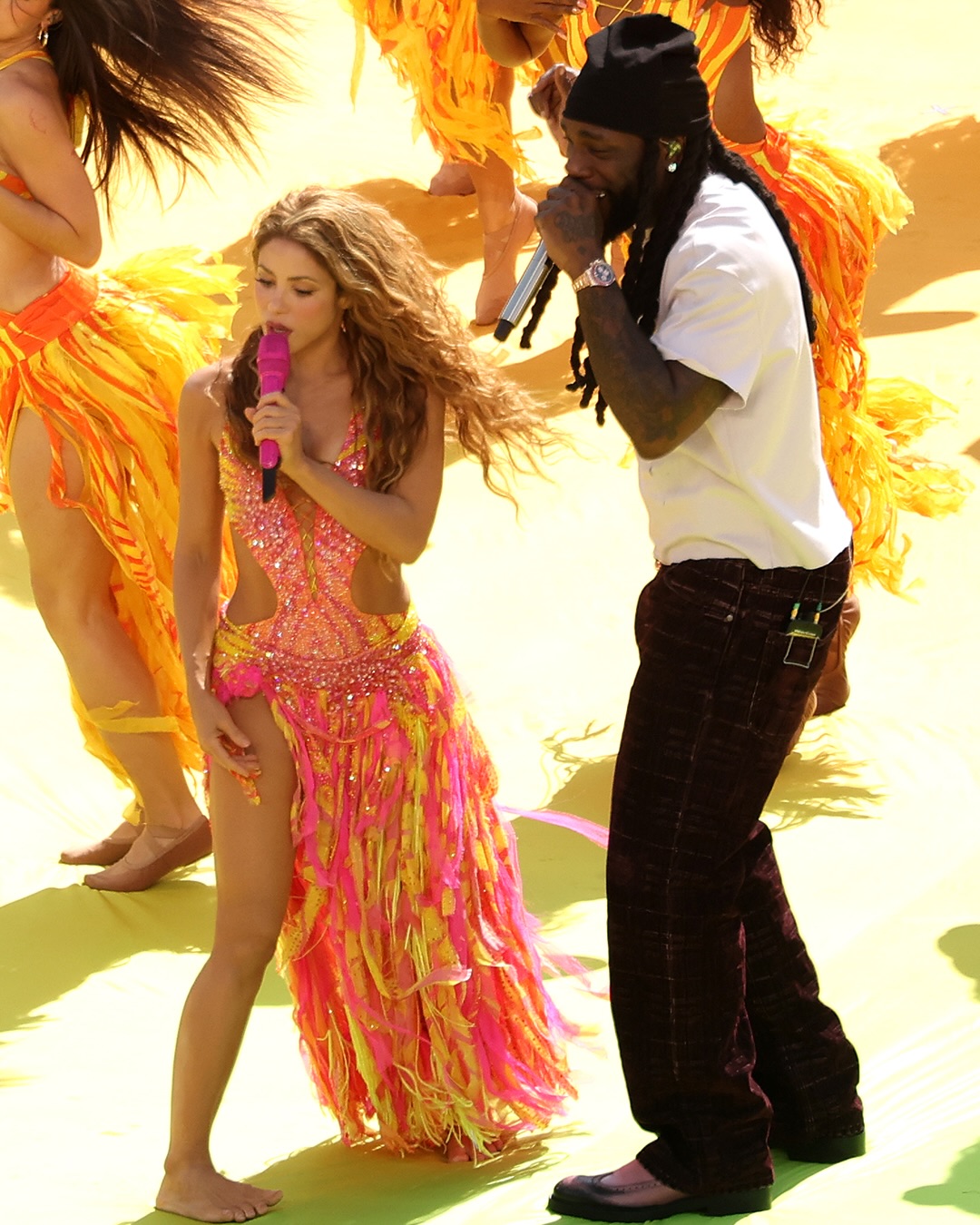 Burna Boy in a white t-shirt, dark trousers, and black beanie sings into a silver microphone beside Shakira performing barefoot on stage.