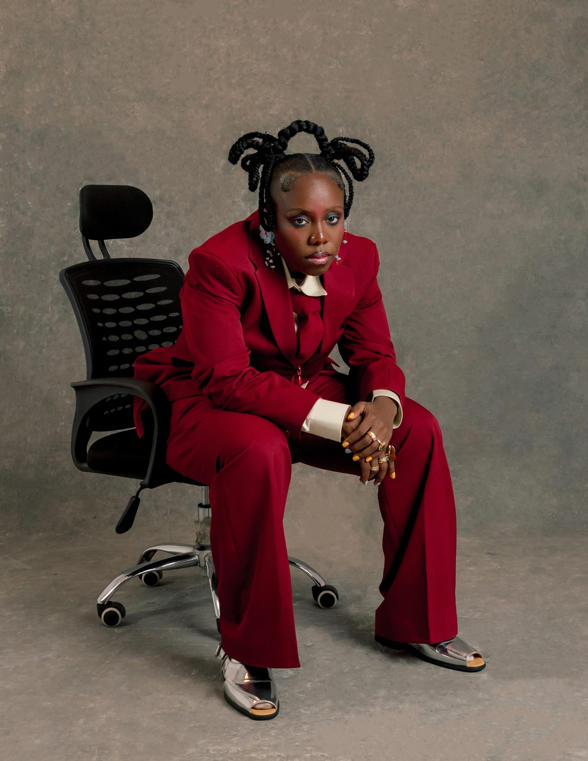 Teni leaning forward seated in an office chair wearing a crimson double-breasted suit, wide-leg trousers, cream shirt with matching red tie, and metallic silver peep-toe slides.