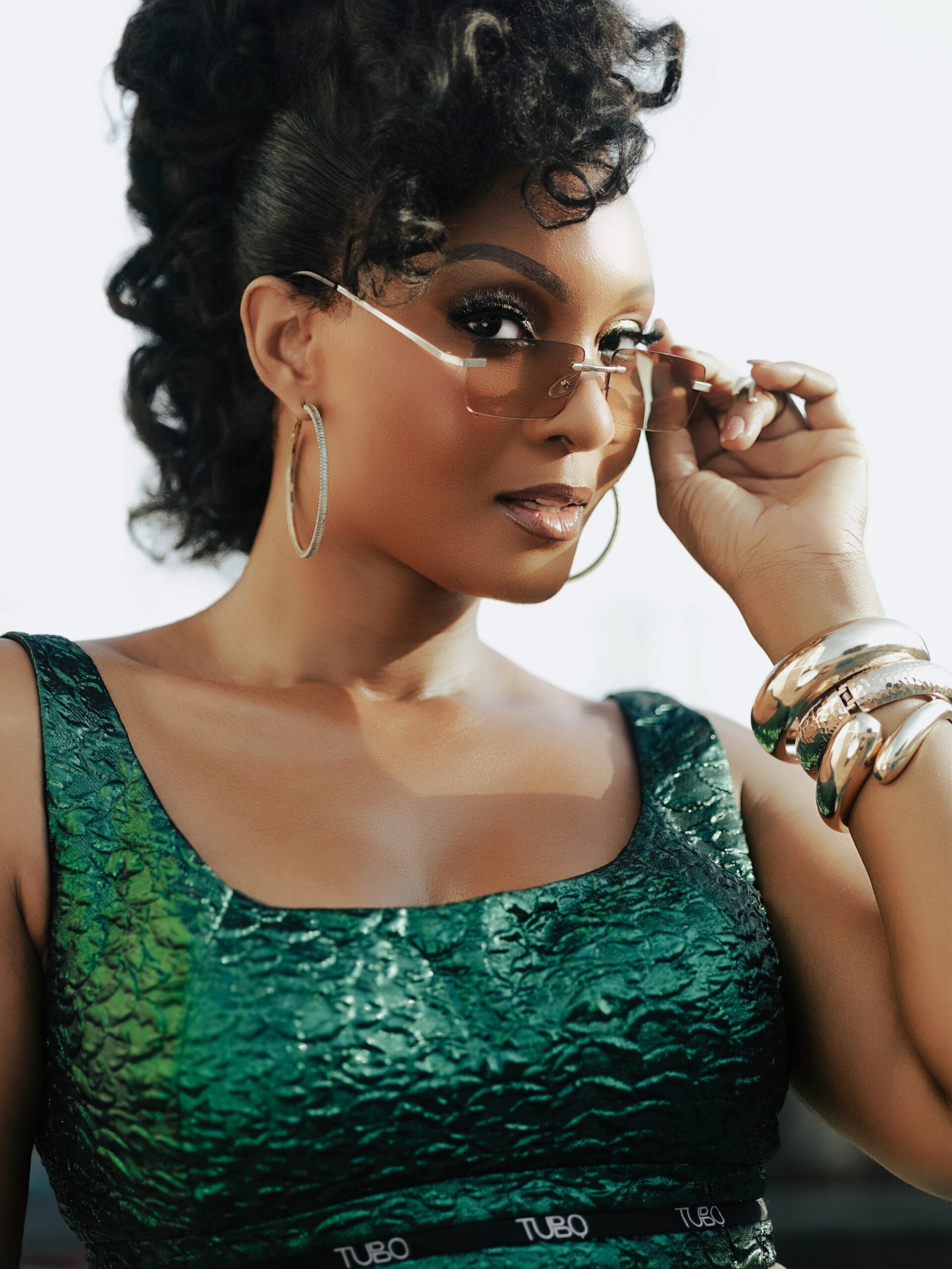 Actress Osas Ighodaro lowering rimless tinted sunglasses, showing dark bronze eyeshadow, oversized hoop earrings, and stacked gold wrist cuffs paired with a green Tubo RTW top.