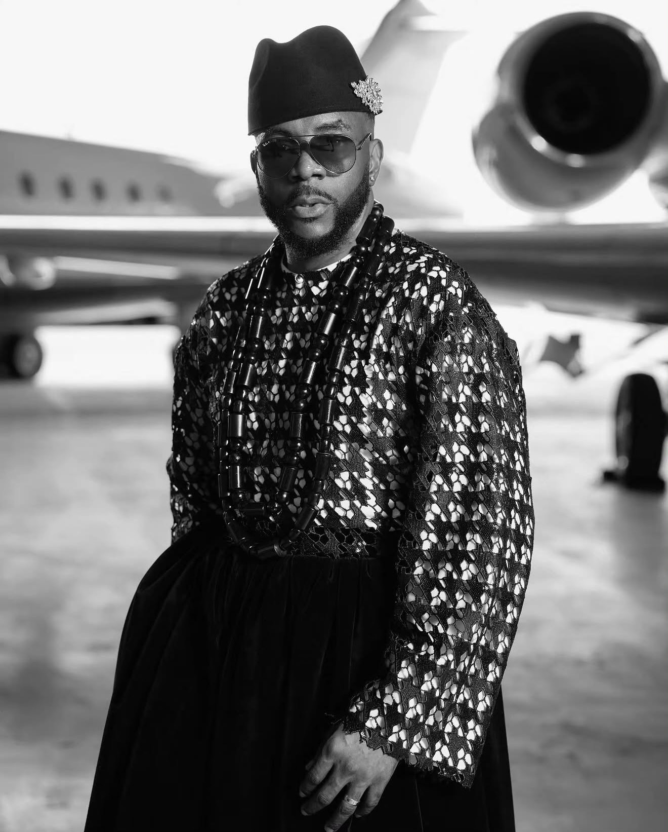 Ebuka Obi-Uchendu wearing a metallic silver and black sequin tunic with heavy black coral beads, traditional cap with crystal brooch, and aviator sunglasses inside an airfield hangar for Big Brother Naija Season 11