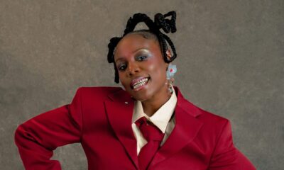 Teni standing with hands on her hips wearing a crimson double-breasted suit, cream shirt, red tie, and showing a silver grillz smile.