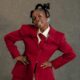 Teni standing with hands on her hips wearing a crimson double-breasted suit, cream shirt, red tie, and showing a silver grillz smile.