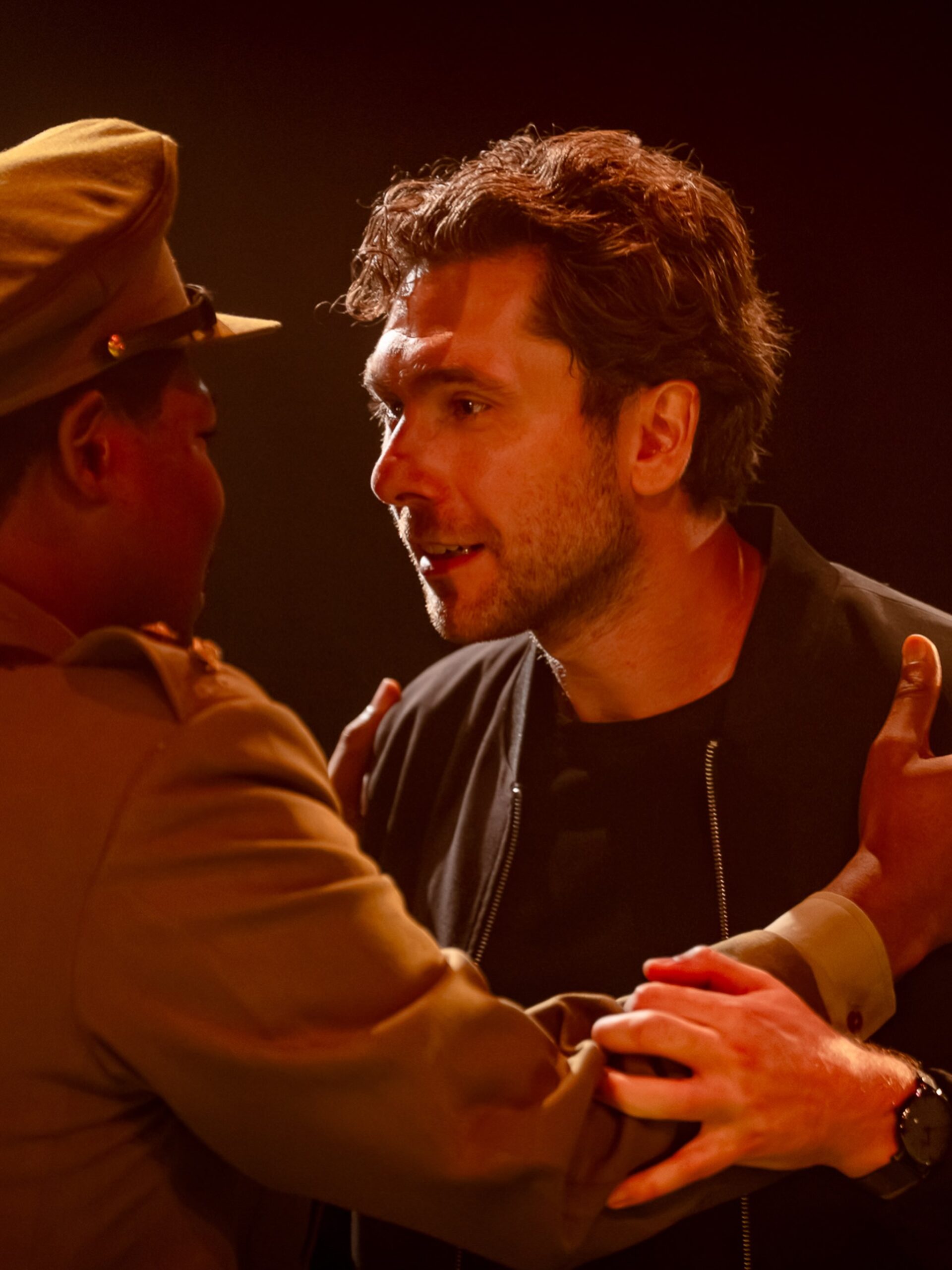 Debo Adedayo seen from behind in military officer uniform gripping the arms of a male actor in dark clothing under dramatic red stage lighting.