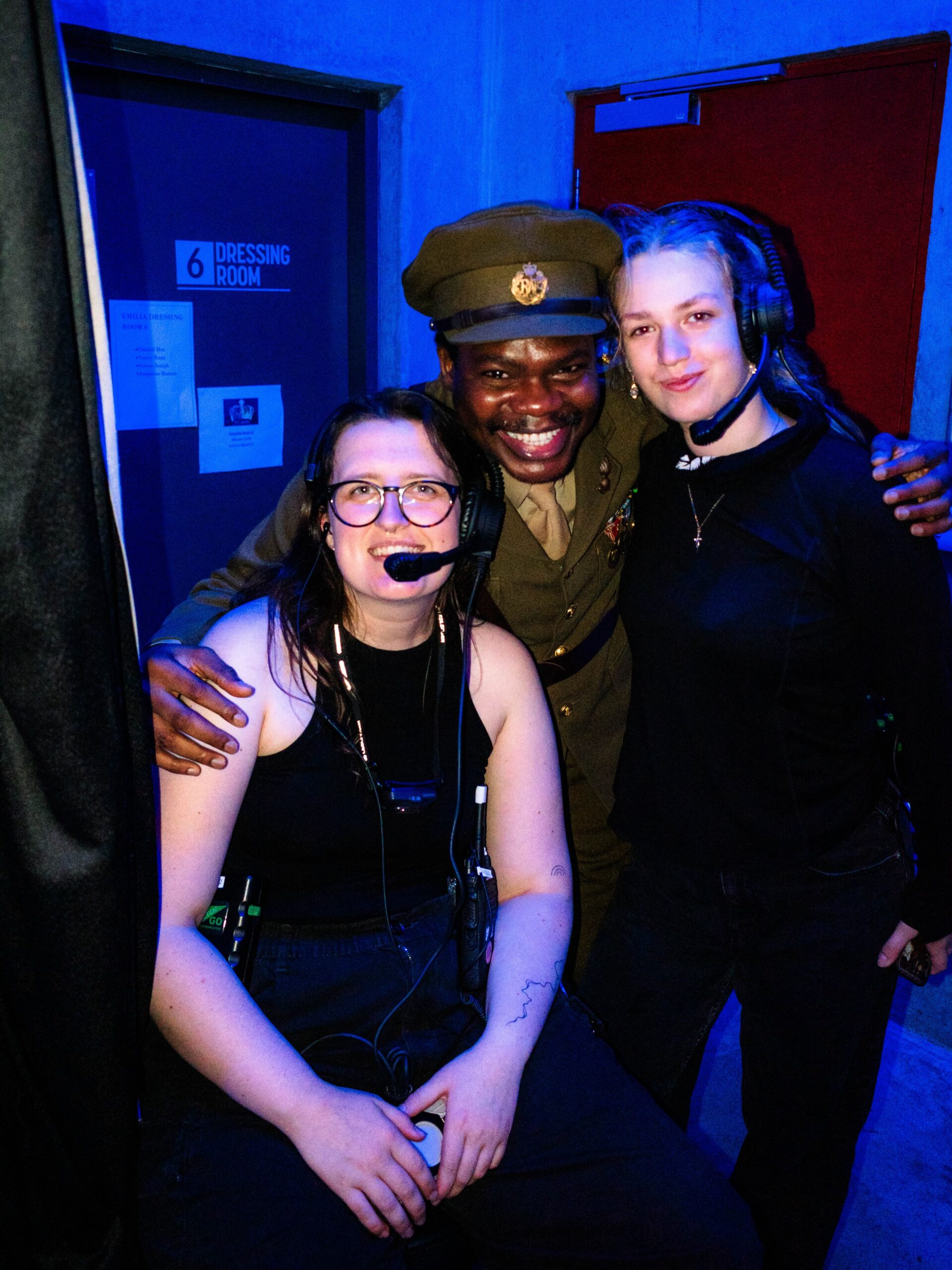 Debo Adedayo smiling in his Lord Commander Thomas Fairfax military costume while standing backstage with two headset-wearing production crew members in London.
