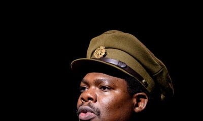 Debo Adedayo wearing a olive-green military tunic with medals, shoulder insignia, matching tie, and officer peaked cap performing in 55 Days at The Backstage Theatre London.
