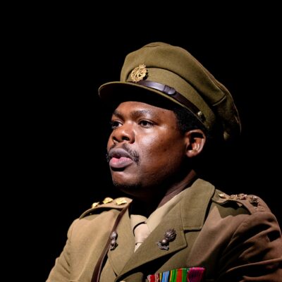 Debo Adedayo wearing a olive-green military tunic with medals, shoulder insignia, matching tie, and officer peaked cap performing in 55 Days at The Backstage Theatre London.