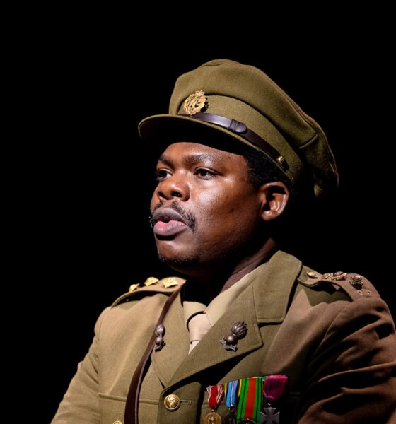 Debo Adedayo wearing a olive-green military tunic with medals, shoulder insignia, matching tie, and officer peaked cap performing in 55 Days at The Backstage Theatre London.