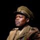 Debo Adedayo wearing a olive-green military tunic with medals, shoulder insignia, matching tie, and officer peaked cap performing in 55 Days at The Backstage Theatre London.