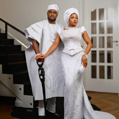 Femi Atere resting his hand on a carved lion cane alongside Veekee James wearing a pearl-beaded white gown and white gele.