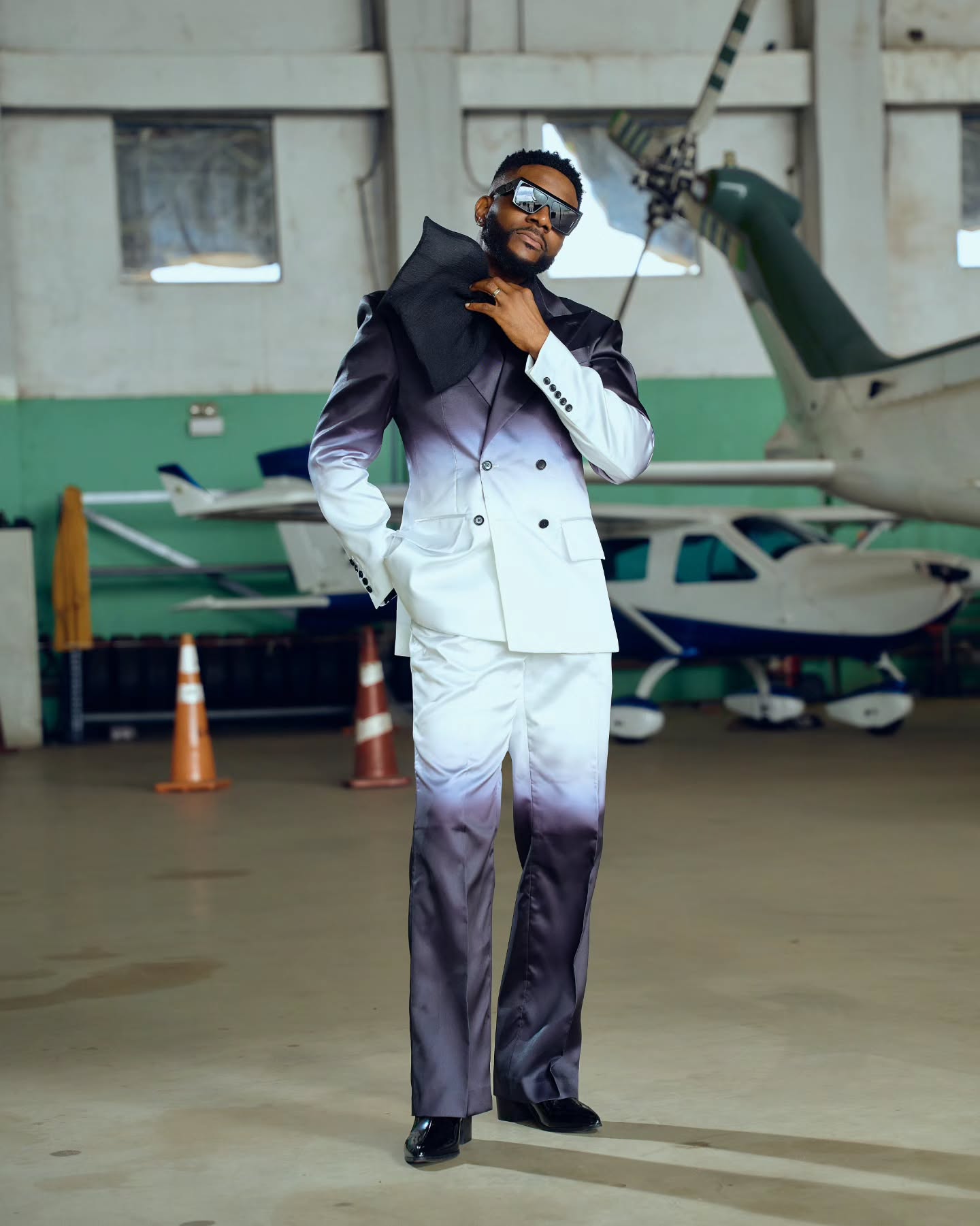 Ebuka Obi-Uchendu wearing a custom ATAFO double-breasted ombré suit in charcoal black and off-white with a large black neck bow, oversized sunglasses, and black patent boots inside an airfield hangar for the Big Brother Naija Season 11 launch.