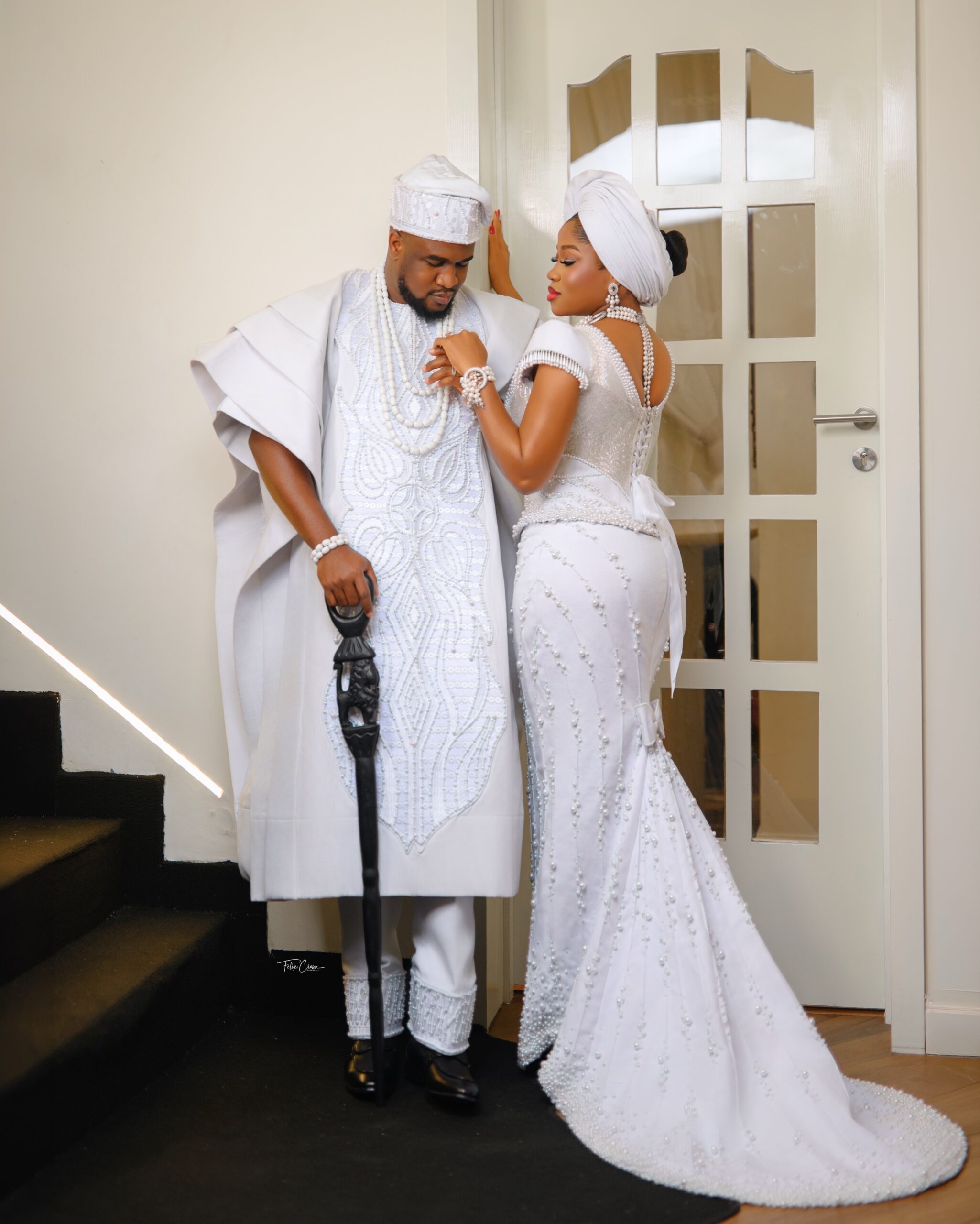 Femi Atere in a multi-layered architectural white Agbada standing beside Veekee James featuring a lace-up corset back and sweeping pearl-strewn train.