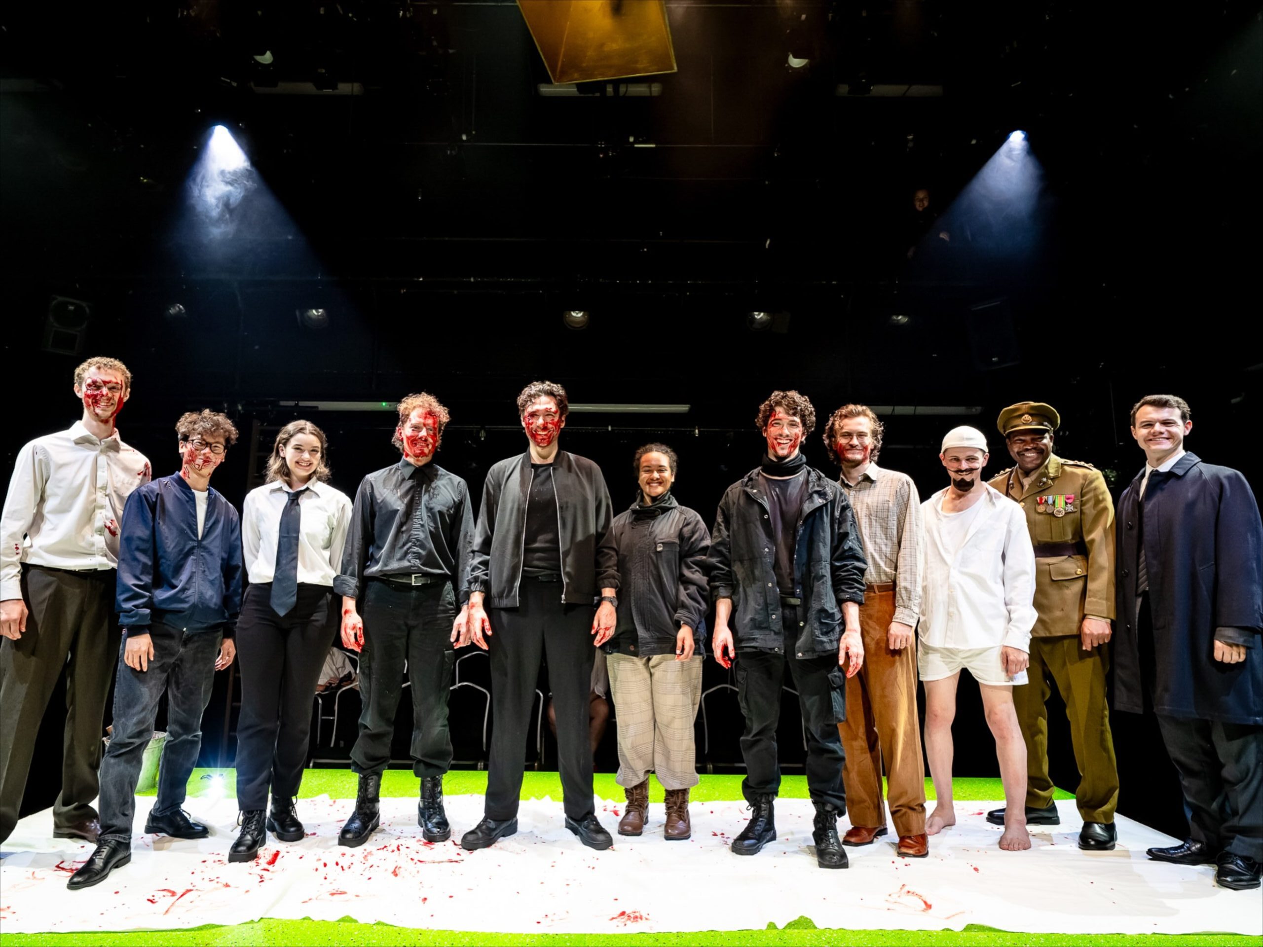 The complete cast of 55 Days standing on stage at Mountview in London, including Debo Adedayo in military costume alongside actors featuring theatrical blood makeup.