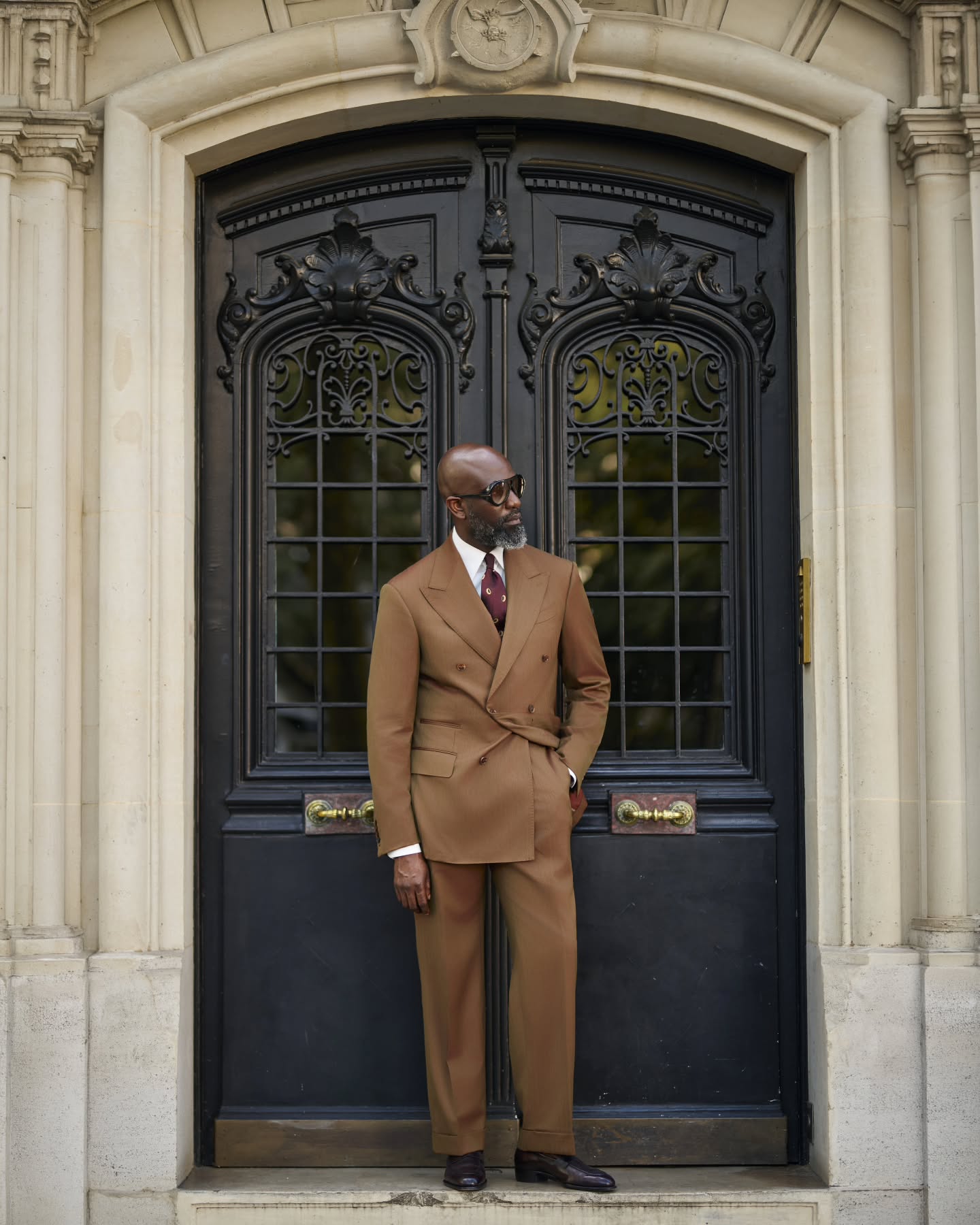 Mai Atafo standing in front of an ornate black door in Paris wearing a fully buttoned tobacco brown double-breasted suit with wide peak lapels, double flapped pockets, a burgundy tie, and dark oxblood loafers.