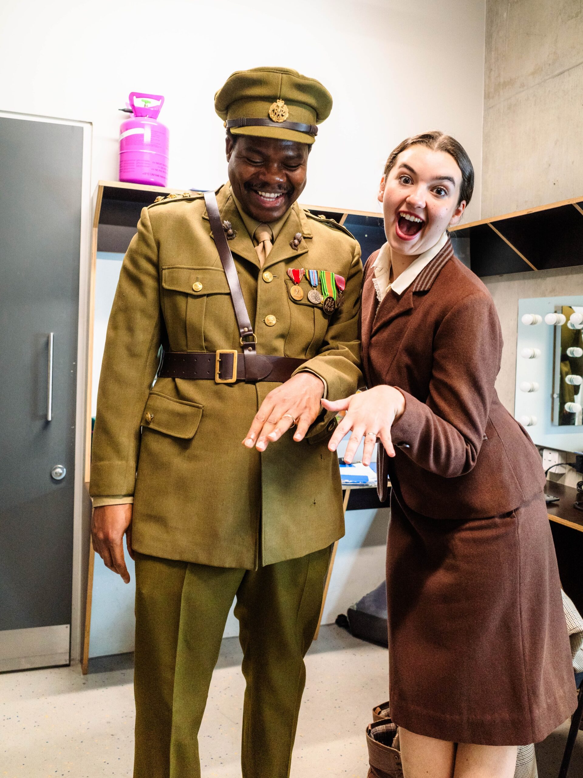 Debo Adedayo and a female co-star in brown tailored period costumes displaying matching prop rings on their hands inside a Mountview dressing room.