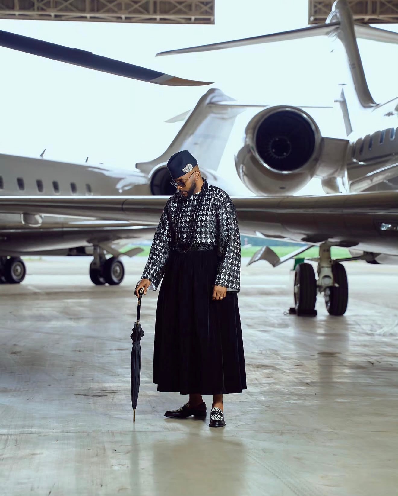 Ebuka Obi-Uchendu holding a black umbrella cane while wearing a metallic sequin lace tunic and floor-length velvet wrapper by ATAFO in an aircraft hangar for Big Brother Naija Season 11.