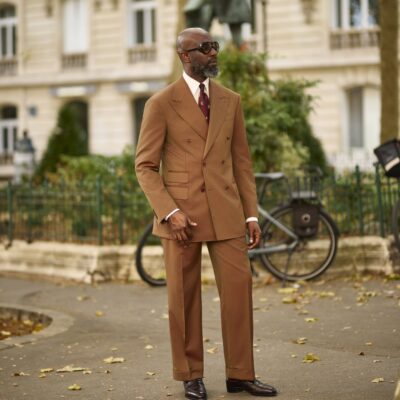 Mai Atafo posing on a Paris street wearing a tobacco brown suit with one hand in his trouser pocket, styled with a white shirt, dimpled burgundy tie, dark aviator sunglasses, and oxblood penny loafers.