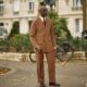 Mai Atafo posing on a Paris street wearing a tobacco brown suit with one hand in his trouser pocket, styled with a white shirt, dimpled burgundy tie, dark aviator sunglasses, and oxblood penny loafers.
