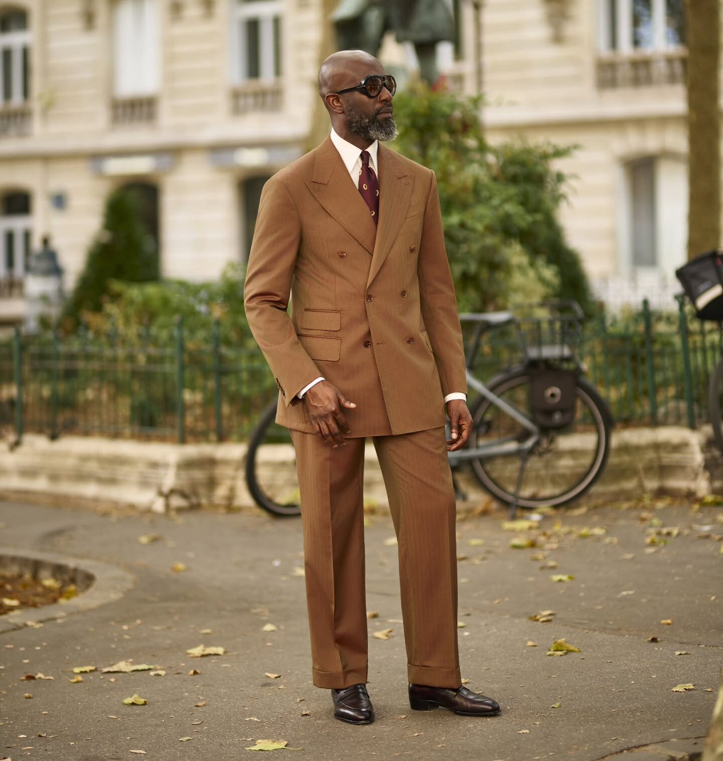 Mai Atafo posing on a Paris street wearing a tobacco brown suit with one hand in his trouser pocket, styled with a white shirt, dimpled burgundy tie, dark aviator sunglasses, and oxblood penny loafers.