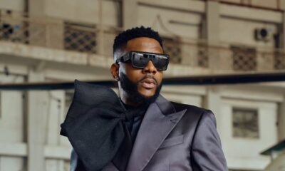 Ebuka Obi-Uchendu in a double-breasted ATAFO jacket transitioning from charcoal black to off-white, styled with a sculptural black lapel bow, peak lapels, and square sunglasses at the Big Brother Naija Season 11 launch.