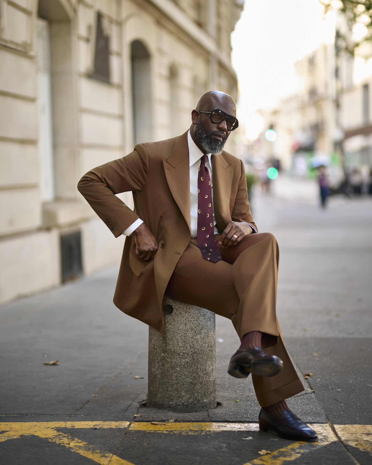 Mai Atafo sitting on a street bollard in Paris wearing a tobacco brown double-breasted suit, crisp white dress shirt, patterned burgundy silk tie, ribbed burgundy socks, dark oxblood leather penny loafers, and aviator sunglasses.