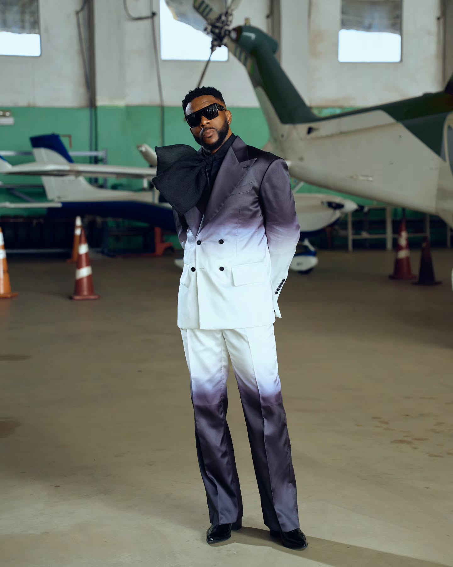 Ebuka Obi-Uchendu in a double-breasted ATAFO jacket transitioning from charcoal black to off-white, styled with a sculptural black lapel bow, peak lapels, and square sunglasses at the Big Brother Naija Season 11 premiere.