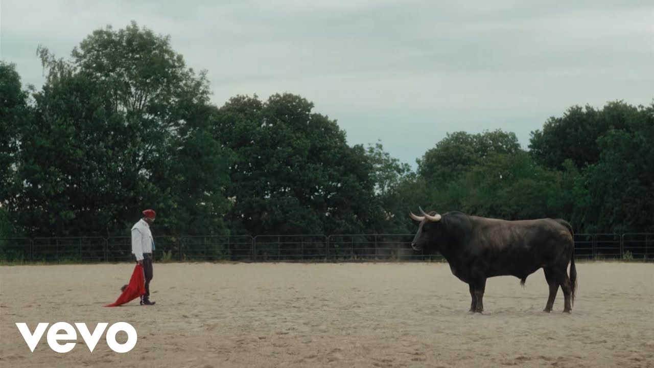 Asake dressed as a matador in a white embellished jacket and red cap, facing a large black bull in an arena.
