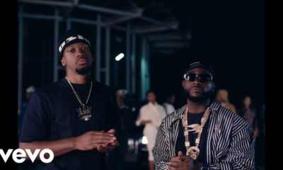 Houston-born singer NO11 in a black tee and beanie standing next to Davido who is wearing a striped shirt, sunglasses, and layered gold chains in the Gimme Dat Ting music video.