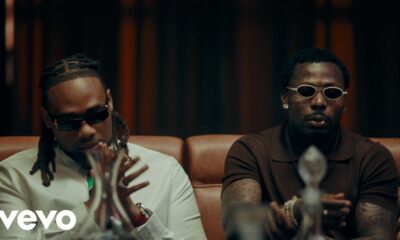 BNXN wearing a white shirt with a colourful striped tie and dark sunglasses seated beside Asake in a brown polo shirt and white oval-framed glasses in the Eja Meja music visualiser.