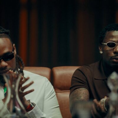 BNXN wearing a white shirt with a colourful striped tie and dark sunglasses seated beside Asake in a brown polo shirt and white oval-framed glasses in the Eja Meja music visualiser.