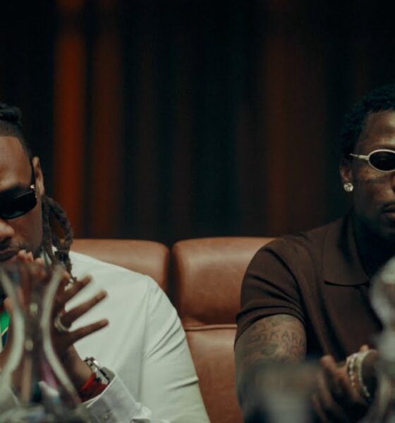 BNXN wearing a white shirt with a colourful striped tie and dark sunglasses seated beside Asake in a brown polo shirt and white oval-framed glasses in the Eja Meja music visualiser.