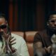 BNXN wearing a white shirt with a colourful striped tie and dark sunglasses seated beside Asake in a brown polo shirt and white oval-framed glasses in the Eja Meja music visualiser.